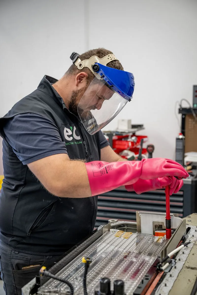 EV Clinic technician in a face shield and class 0 high-voltage gloves, working inside an opened battery module.
